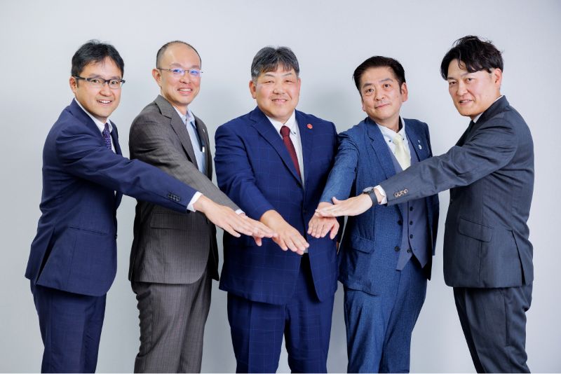 写真：令和7年度支援先企業の皆さんが笑顔で手を組むようす